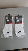 Two pairs of white Nike socks, new, each individually packaged with black Nike label and hangers on a gray surface.