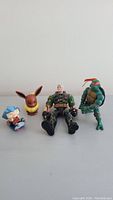 Toy figures arranged against a white background including a Teenage Mutant Ninja Turtle, Eevee, seated blue-haired figure, and military action figure.