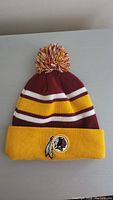 Front view of the knit pom-pom beanie hat showing the Washington Redskins logo patch on the yellow cuff and maroon, yellow, white color bands and pom-pom.