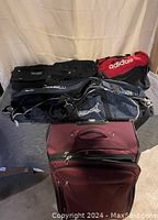 Six assorted bags including sports duffles and rolling luggage shown together on floor against beige curtain background