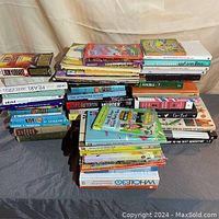 Photo showing large stacks of assorted books in various sizes and genres, stacked horizontally on a black surface against a beige curtain background.