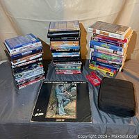 Photo of multiple stacks of DVDs, vinyl record, audio cassettes, and portable DVD player in black case on fabric-covered table.