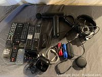Image showing multiple remote controls, a handheld massage gun, two types of headphones including earbuds and over-ear wired headphones, a Fitbit wristband, red and blue flashlights, charging cables, and a small black wireless speaker with strap.