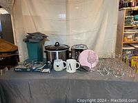 Various kitchen items including stainless steel Crock-Pot, Black & Decker toaster, glassware, mugs, and ravioli maker on a table with fabric placemats stacked in the background.
