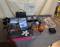 Lot contents displayed on a table including earbuds in boxes, jewelry, hair dryer, curling iron, suction mounts, magnifying sheet, and various small items.