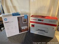 Front view of Canon PIXMA MG2522 box and HP ENVY 6055 box wrapped in plastic on a table.