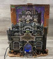 Front view of the Lemax Dr. Stretch N. Pull's Torture Factory with original box behind, showing detailed Gothic-style structure, lighting, and two grim reaper figures at the doorway.