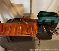 Wide view showing all items in the lot including the Iworx plow attachment, Makita bag, black plastic box, and tools on the table