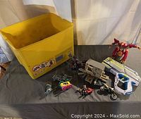 Yellow storage bin with various toys on a dark surface including action figures, LEGO pieces, a toy gun, and a toy motorcycle.