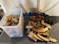 Wooden train track pieces inside a plastic bin and spread out on a surface including straight, curved, and switch pieces plus a wooden bridge and accessories.