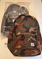 Two Herschel backpacks shown: one camouflage and one grey in plastic packaging, front view.