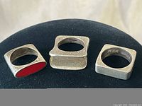 Three modern square rings displayed on black background showing different designs: red coral inlay, solid smooth, and folded metal detail.