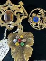 Three vintage brooches arranged on a black background, showing detail of a leaf-shaped brooch with colored rhinestones, round brass floral brooch with blue stone, and brass brooch with amber-colored stone.