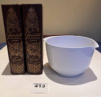 Two Gourmet Cookbook volumes I and II 1959 editions standing upright with brown hardcovers and gold ornate embossing, next to a white Rosti bowl with a pouring spout.