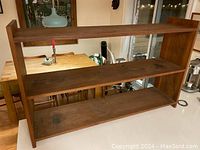 Front view of teak wood shelving unit with three shelves and lower shelf cross support. Shows some staining and wood grain detail.