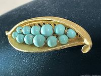 Photo shows front side of gold-tone leaf-shaped brooch with turquoise beads in two rows inside.