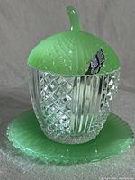 Full view of the vintage sugar bowl with its matching lid and saucer. Crystal glass body with textured diamond and vertical patterns. Jadeite green lid with handle and textured pattern. Jadeite green saucer with scalloped edges.