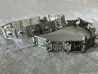 Two vintage sterling silver bracelets side by side: ornate filigree panel bracelet and zodiac sign panel bracelet showing their overall design and clasp closures.