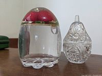 Clear acrylic or perspex sugar bowl or jelly jar with a red raspberry top lid and green leaf accents, next to a pear-shaped cut glass trinket box with intricate patterning and a lid.