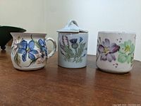 Front view of three ceramic items: two floral mugs and a lidded jar on a wooden surface, white background.