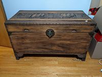 Front view of the wooden chest showing carved details, metal lock, and feet, placed on a wood floor.