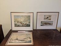 Three framed prints on a dark wood table against a white wall, featuring winter scenes by Cornelius Krieghoff