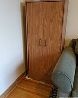 Tall brown laminate cabinet with wood grain finish, two metal handles, standing next to a couch on hardwood floor.