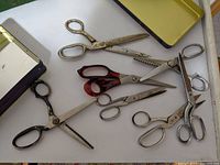 Seven vintage sewing scissors including two pinking shears on a white surface near open metal boxes.