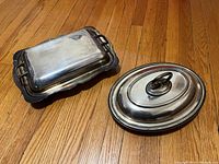 Two silver plated serving dishes with lids shown side by side on hardwood floor. One rectangular with handles and black tray, the other oval with domed lid.