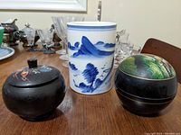 Three items on a table: a white ceramic brush pot with blue landscape painting, a black lacquer box with floral and bird design lid, and a larger black lacquer nesting box with a painted nature scene lid.