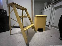 Side angle view of yellow painted vintage metal step ladder and foot-operated waste can side by side on floor.