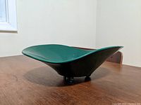 Side angle view showing the wavy, undulating edges and smooth glazed green surface. The dish sits elevated on three small round ball feet.