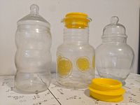 Lot showing vintage Pyrex lemonade jug with original yellow lid and bonus yellow lid, flanked by two clear curvy glass candy jars with lids.