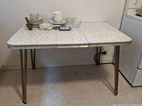 Vintage retro style rectangular table with chrome edges and legs under artificial indoor lighting, showing the patterned cream Formica-type table top and chrome legs.