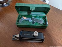 Vintage Singer buttonholer attachment in front of open green case containing buttonhole templates and parts.