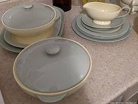 Overall view of the covered vegetable dishes with lids, serving plates stacked, and smaller plates visible beneath, all in muted green with yellow interiors.