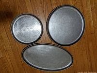 Three large stainless steel trays shown from above on wooden floor, two circular with rope edge detail, one oval shape