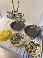 Three ceramic Anthropologie plates with yellow rims and colorful fruit designs, two metal 9 inch bakers pans, and bottom of one plate showing branding and date '2021 Nathalie Lété for Anthropologie'.