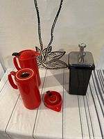 Top view of two red coffee thermal dispensers and one black ceramic antique dispenser with pump top on white cloth surface.
