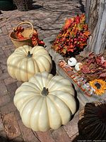 Photo showing 2 large off-white plastic pumpkins, fall leaf wreath with berries, woven basket with orange ribbons, wooden 'Give Thanks' sign, and pumpkin candle on outdoor brick surface.