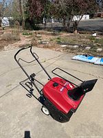 Left-front angle showing red housing, steel handle, and black discharge chute.