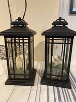 Pair of black metal Luminara candle lanterns with visible faux candles and artificial greenery inside, placed on striped cloth.