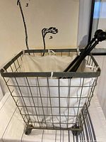 Metal rolling laundry cart with white cloth insert liner, viewed at an angle shows the metal frame, cloth liner, and caster wheels on a white surface.
