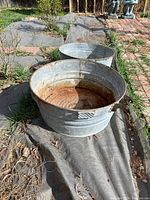 Outdoor photo of two round vintage wash bins with handles, showing rust and wear on metal surfaces, placed on ground covering near brick pathway.