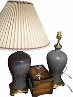 Two grey ceramic lamps with brass bases. One lamp has a beige pleated shade, the other lamp is missing a shade. Lamps are glossy and bulb-shaped.