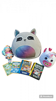 Photo shows a collection of kids toys and books arranged together including a large grey Squishmallow raccoon plush, a pastel-colored fairy doll, four Jumbo Pokemon cards laid out, three Captain Underpants books, and a Squishmallow-style unicorn doll head.