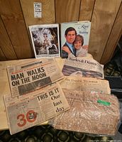 Overview photograph showing multiple vintage newspapers and royal wedding publications on wooden floor surface with wood panel wall background.