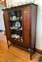 Full view of antique wood china cabinet showing three tiers, glass front door with inlaid wood, and contents inside (not included). Cabinet stands on four legs with stretch bar.