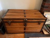 Front view of antique wooden trunk showing wooden slats, metal edges, two latches, and central lock plate with decorative circular metal accent.