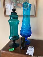 Two glass vases and one glass jar with lid on wooden surface with framed artwork in background.